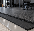 Black rubber gym flooring tiles with a speckled texture, installed on a gray concrete base in a gym setting.