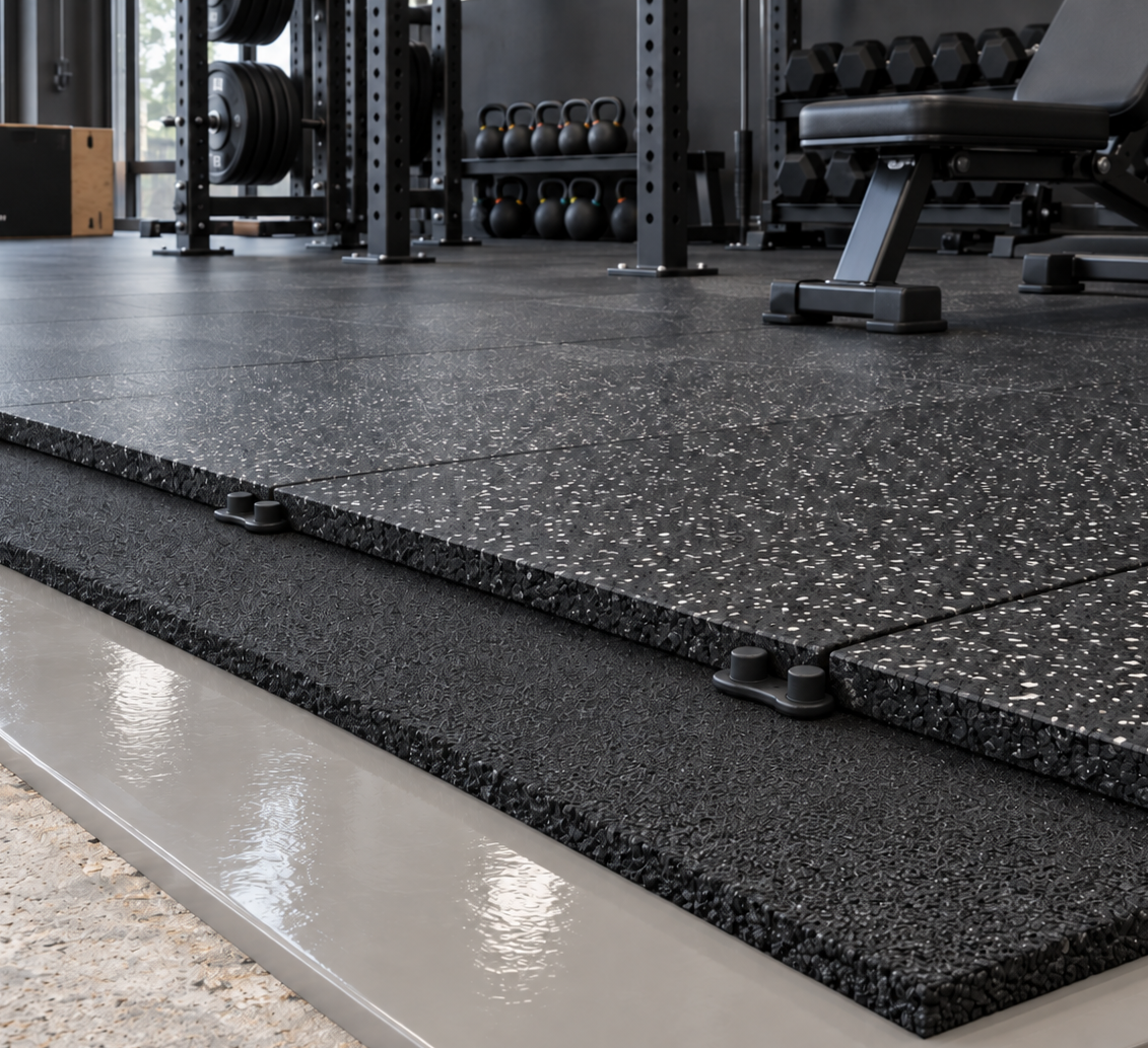 Black rubber gym flooring tiles with a speckled texture, installed on a gray concrete base in a gym setting.