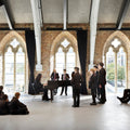 A group of people in formal black clothing gather in a large, bright hall with arched windows, brick walls, and exposed white beams. A woman sits at a black grand piano while several men stand nearby, and a seated audience watches from the floor.