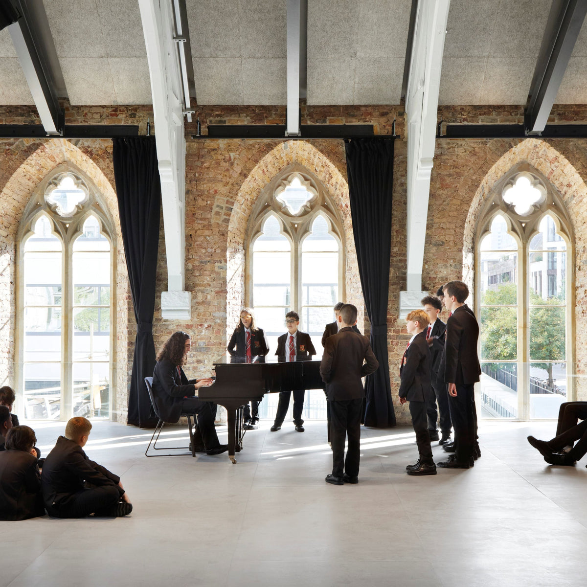 A group of people in formal black clothing gather in a large, bright hall with arched windows, brick walls, and exposed white beams. A woman sits at a black grand piano while several men stand nearby, and a seated audience watches from the floor.